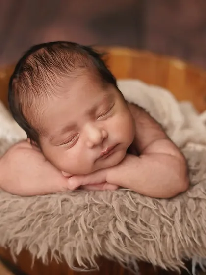 A close-up of a newborn sleeping peacefully in a wooden bucket, resting his head on his arms. The soft textures and warm tones create a very cozy and rustic feel.