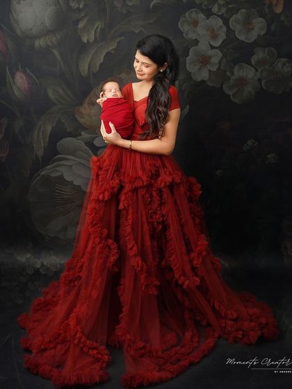 A serene portrait of a mother and her newborn. Dressed in a gorgeous red gown, she holds her sleeping baby, creating a peaceful and elegant image against a classic dark backdrop.