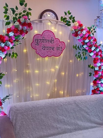 A simple yet elegant baby shower decoration perfect for a home setting. It features a floral arch over the main sofa with a 'Kunitari Yenar Ga!' sign against a fairy-lit curtain.
