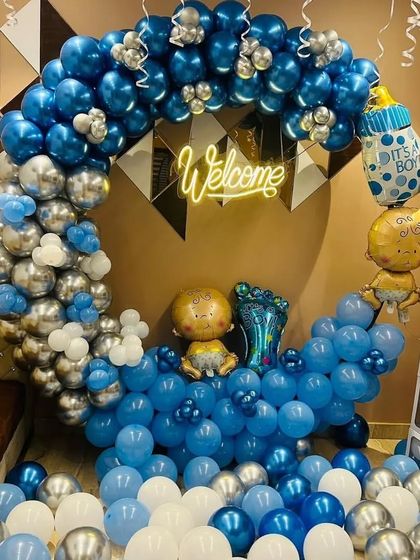 A grand welcome setup for a baby boy in a living room. This features a large crescent-shaped balloon arch with blue and silver chrome balloons, a "Welcome" neon sign, and multiple baby-themed foil balloons.