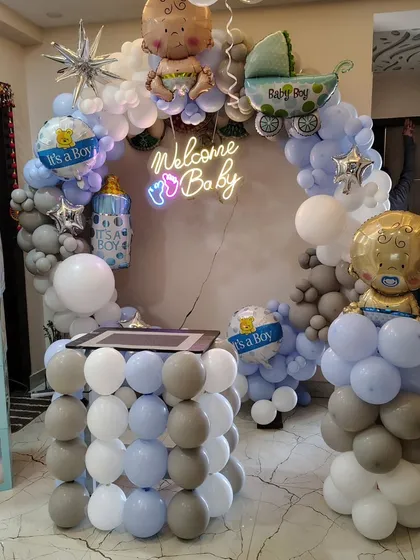 A "Welcome Baby" boy decoration with a blue, grey, and white balloon ring, "BABY" blocks, and a neon sign.