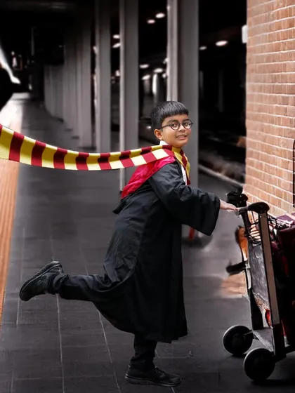 A magical Harry Potter themed photoshoot featuring a young boy in a Gryffindor robe pushing a luggage trolley towards a Platform 9¾ sign on a brick wall.