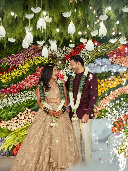 This reception stage features a unique, artistic wall of flowers arranged in colorful waves, with hanging lights that look like floating buds.
