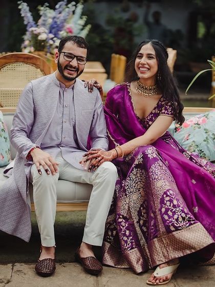 A couple in beautiful purple and lavender outfits sits on a vintage sofa, a perfect portrait from their Mehendi or Sangeet.