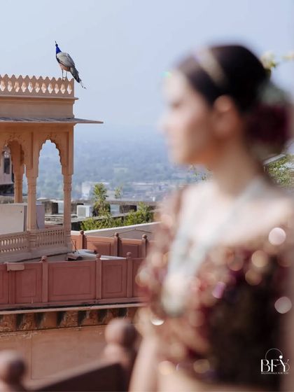 An artistic shot of a bride out of focus, with a peacock perched on the fort in the background.