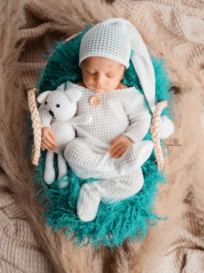 A sleeping newborn in a cozy, textured white romper and matching hat, holding a small knitted toy. This boho-inspired shot uses natural textures like pampas grass for a warm, earthy feel.