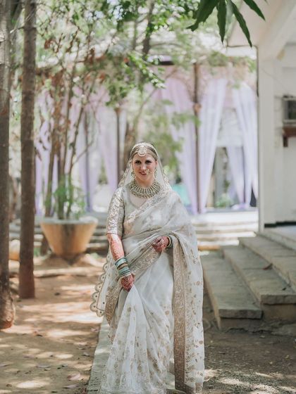 A bride in a white saree, her face framed by her veil, stands in the garden, a picture of ethereal beauty.