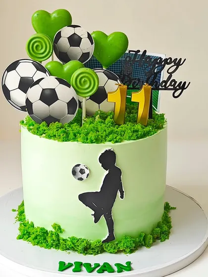 A vibrant green football-themed cake for an 11th birthday. The design features a player silhouette, soccer balls, and a "grass" border made from cake crumbs.