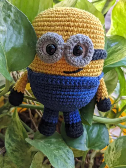 The crochet Minion doll peeking out from behind some green leaves.