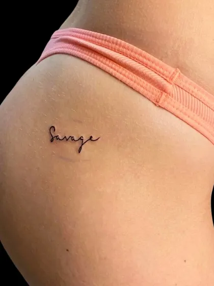 A "Savage" script tattoo on the hip. This is a bold and confident statement piece, done in a clean and simple font.