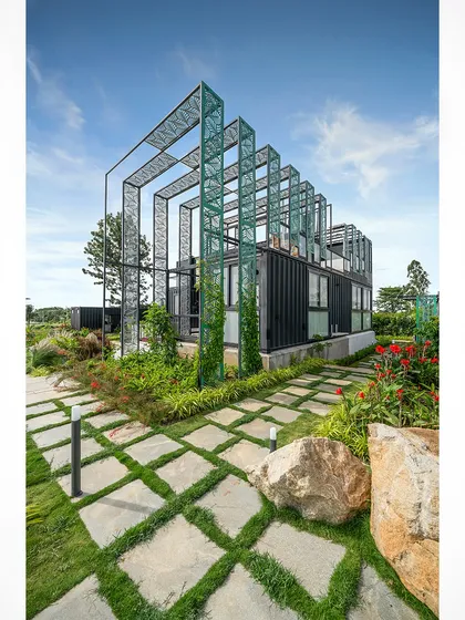 A daytime view of the container home, showing the green perforated metal structure that adds a sculptural element to the building. The surrounding greenery and stone pathway integrate the home into its landscape.