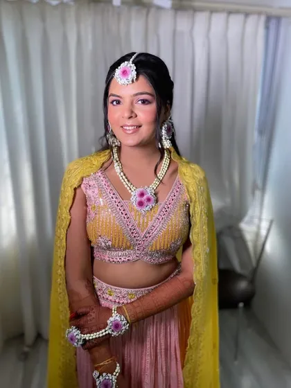 A pretty and fresh look for a haldi ceremony. The makeup is minimal and pink-toned, and the hair is styled in a half-updo with a beautiful floral and pearl hairpiece.