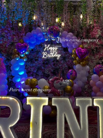 A close-up of a unicorn-themed birthday backdrop. The "Happy Birthday" neon sign and heart-shaped foil balloons add a magical glow to the floral and balloon arrangement.