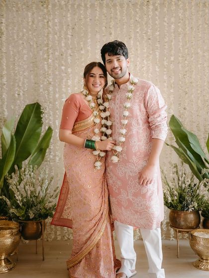 A classic portrait of the couple after their signing ceremony. The decor features a wall of white floral strings, large banana leaves, and traditional brass urlis.