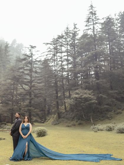 A majestic wide-angle shot of the couple in a misty forest clearing. The bride's long, flowing gown creates a stunning visual against the tall pine trees, perfect for a dramatic pre-wedding portrait.