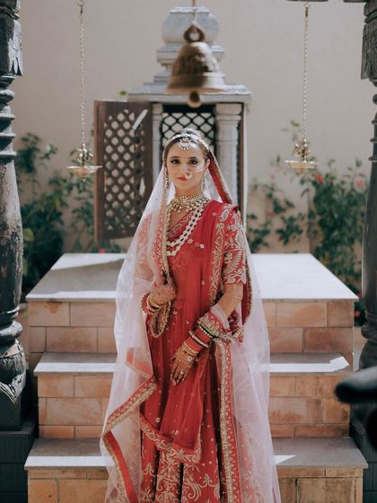 The bride stands before a temple-like structure, her red lehenga a striking contrast to the stone. This portrait has a regal and almost divine quality.