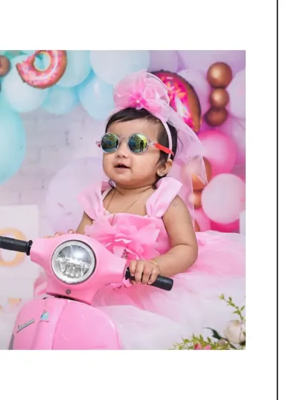 A close-up of the stylish baby girl on her pink scooter, smiling for the camera. The donut and balloon background adds to the sweet, celebratory feel of this candy-themed session.