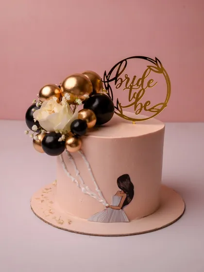 A chic and modern cake for a bride-to-be. This tall, single-tier cake has a smooth pale pink finish, a hand-painted illustration of a bride, and a stylish arrangement of black, gold, and white spheres.
