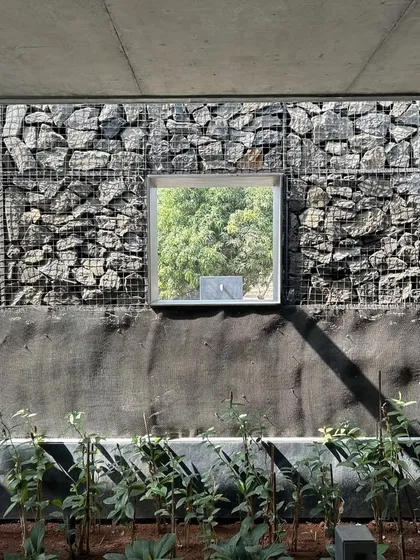 A moment of quiet composition at the Alibaug residence. A perfectly square window is set within the rugged texture of a gabion wall, framing a view of the green foliage outside like a living painting.