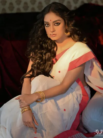 A full portrait from the same series, with the model seated in a classic white and red saree. The moody lighting and vintage wallpaper create a nostalgic, almost cinematic atmosphere.