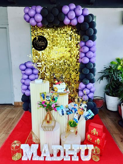 A stylish birthday setup with a gold shimmer wall, framed by a purple and black balloon arch. Personalized elements like photos make the celebration extra special.
