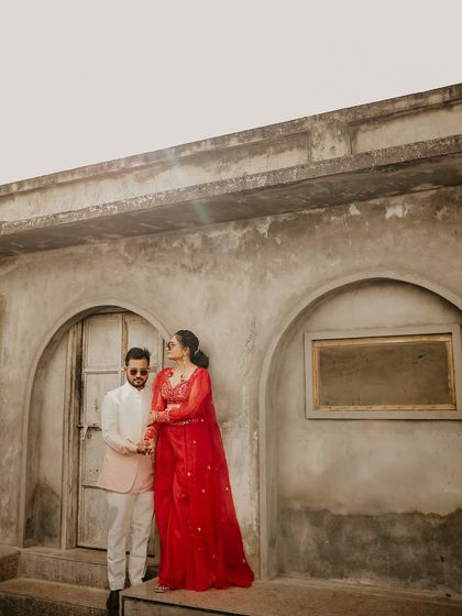 The couple stands against a rustic, textured wall with arched doorways. Their modern yet traditional styling creates a fashionable statement in this pre-wedding portrait.
