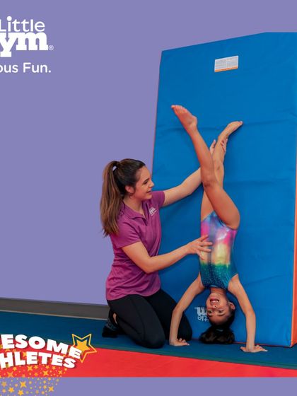 An instructor helps a young 'Awesome Athlete' with a handstand against a soft mat. This technique allows kids to get comfortable being upside down while building the necessary arm and shoulder strength.