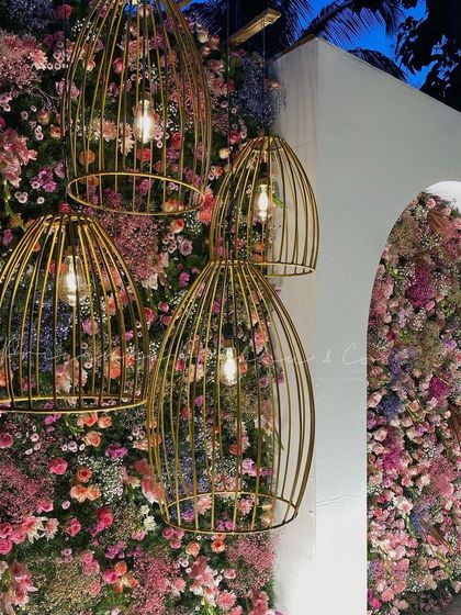 A close-up of the reception stage details. The combination of a dense floral wall with hanging gold birdcage lights creates a beautiful texture and adds a warm, ambient glow to the evening.