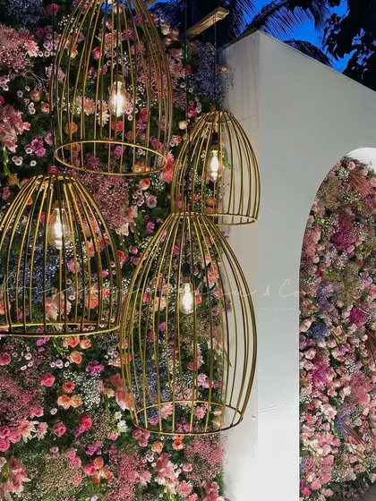 A close-up of the reception stage details. The combination of a dense floral wall with hanging gold birdcage lights creates a beautiful texture and adds a warm, ambient glow to the evening.