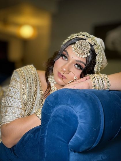 A beautiful bride in a white and gold lehenga, looking relaxed and happy on a blue couch. Her makeup is soft and glamorous, with wavy hair.