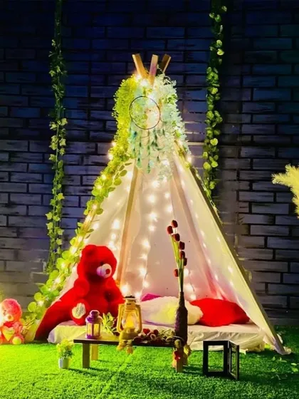A bohemian-style romantic picnic setup at night. A teepee tent is decorated with fairy lights and greenery, creating a magical and intimate setting for two.