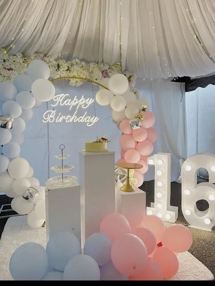 A dreamy sweet 16 birthday party decoration. This setup features a pastel blue and pink balloon arch against a white curtain backdrop, with large light-up numbers for the age.