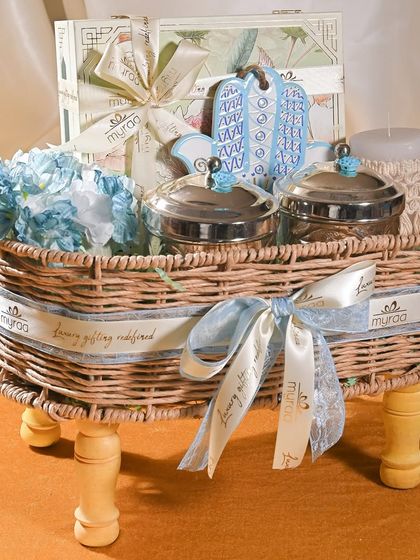 A classic baby boy announcement hamper in a wicker basket on legs. It includes silver-plated jars, a scented candle, and blue floral accents, all tied together with a custom Myraa ribbon.