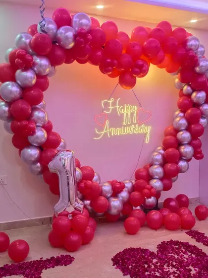 Apni pehli anniversary ko khaas banane ke liye humne yeh red aur silver chrome balloons ka heart shape frame taiyar kiya hai. Beech mein 'Happy Anniversary' ka neon sign iski sundarta aur badha raha hai.