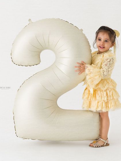 A classic portrait of a little girl proudly standing by her number '2' birthday balloon, dressed in a beautiful yellow lace dress.