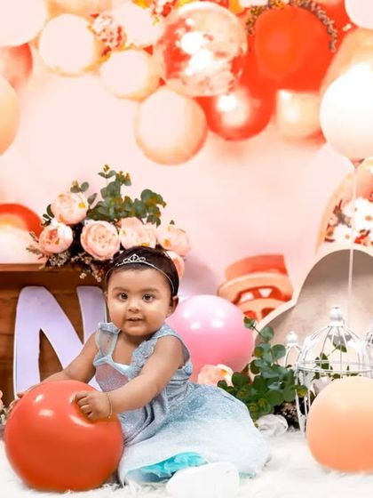 A beautiful first birthday setup with a cascade of peach and white balloons. The little princess is playing with a ball, enjoying her special day.