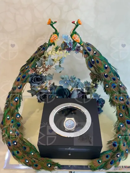 A gift that is both grand and graceful. This luxury watch is presented with two magnificent peacock figurines, their feathers creating a dramatic and opulent frame for a truly special wedding present.