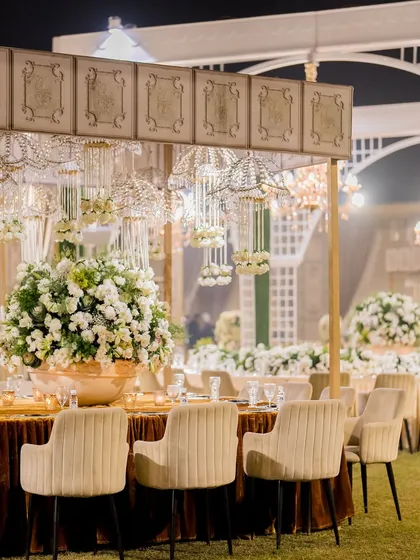 An elegant outdoor dining setup with velvet chairs, gold-rimmed tables, and hanging crystal and floral chandeliers.
