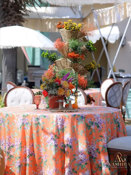 A vibrant tablescape with a floral-printed orange tablecloth, classic chairs, and a whimsical, tiered centerpiece made of woven baskets and colorful flowers.