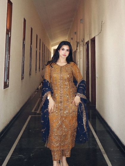 A direct, full-length shot of a mustard and blue ethnic suit by Asim Jofa. The long corridor provides a clean, symmetrical backdrop that puts the entire focus on the detailed craftsmanship of the wedding wear.