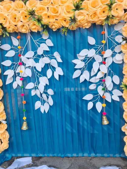For a festive pooja backdrop, I combined a bright yellow artificial rose frame with a rich blue fabric curtain. The design is accented with decorative white leaf hangings and small bells for a unique and modern look.
