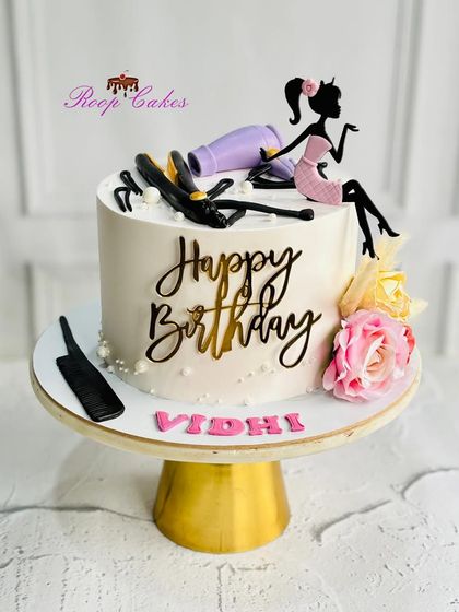 A chic and glamorous birthday cake for a hairstylist. The design features edible salon tools like a hairdryer and straightener, along with a stylish silhouette topper.