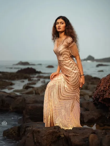 A full-length shot of the model standing on the rocks, with the ocean in the background. The glamorous gown looks stunning against the rugged, natural beauty of the coastline.