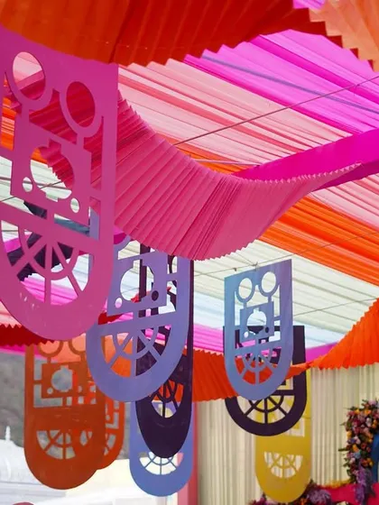 The ceiling decor for the colorful Haldi, featuring drapes in shades of pink, orange, and blue, with decorative cut-outs hanging down. This creates a festive, tent-like feel over the celebration.