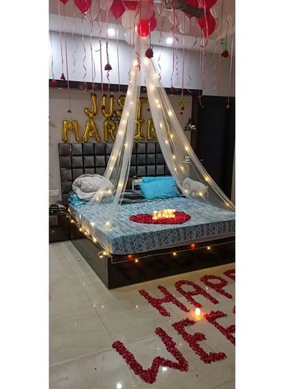 A full view of a "Just Married" bedroom decoration. The design includes a fairy light canopy, "Just Married" foil balloons, and a "Happy Wedding" message written with rose petals on the floor.