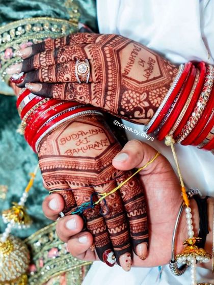 A close-up of the bride and groom's hands, showing the personalized henna stain. The design includes text that is special to the family.
