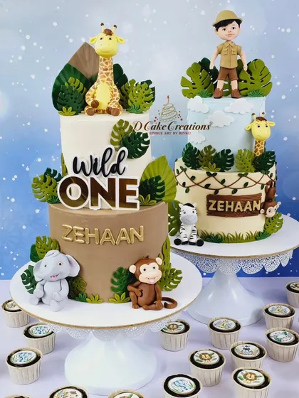 The two stunning jungle-themed cakes for Zehaan's first birthday. One features a "Wild One" sign with cute animals, and the other shows a little explorer among the wildlife.