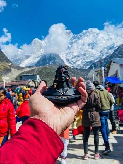Holding a miniature idol of Lord Shiva with the actual Kedarnath temple and snow-capped mountains in the background, connecting the divine symbol with its sacred home.