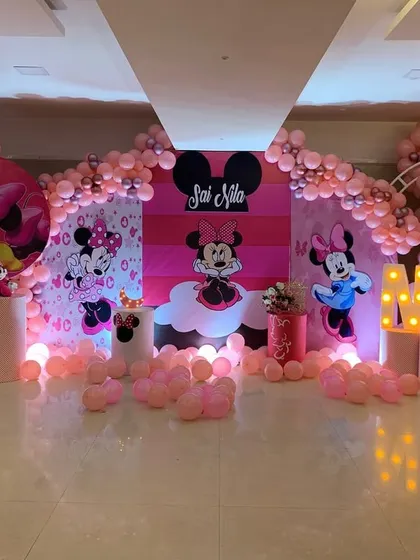 A full view of a Minnie Mouse themed first birthday party. The all pink decor features multiple backdrops with Minnie, balloon arches, and light up initials for a fun and immersive experience.