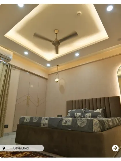 A sophisticated bedroom at Saya Gold with a tall, upholstered headboard and a coffered false ceiling. The neutral color palette creates a calm and restful environment.